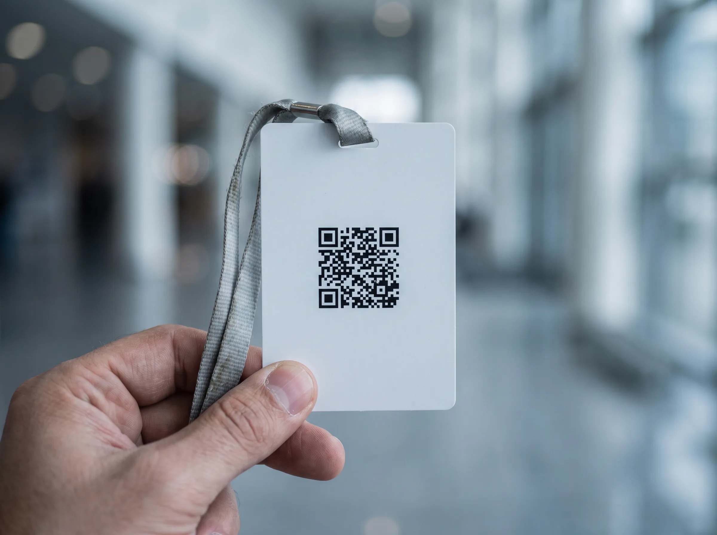A hand holding a plain white conference badge on a grey lanyard, carrying a QR code and nothing else, with the venue blurred behind it.