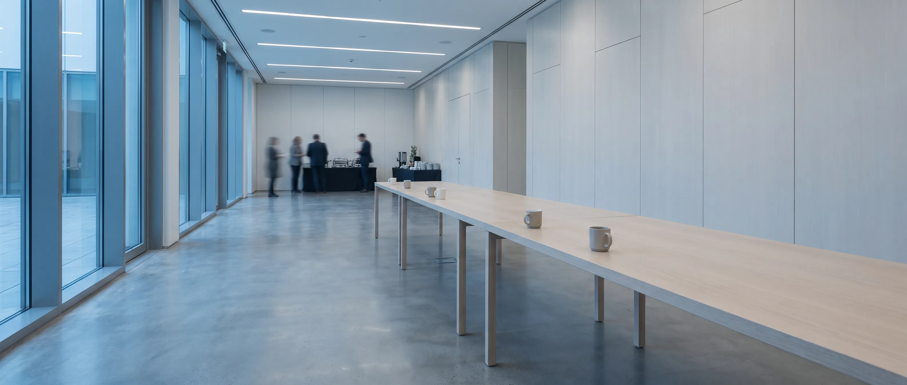 A modern conference hall between sessions: pale walls, a long table with cups left on it, daylight through a glazed wall, and a few people talking at the far end.