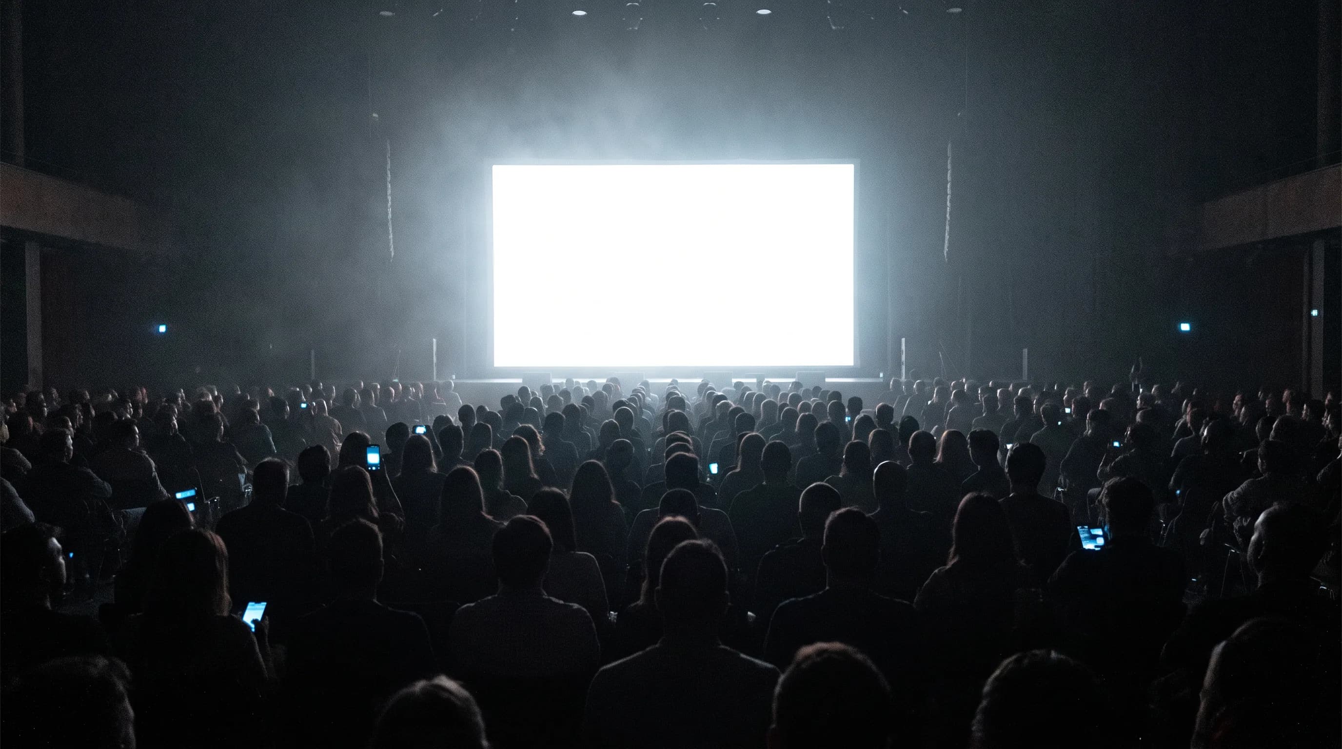 The back of a darkened auditorium during a talk: rows of silhouetted heads, a blank projection screen throwing light over them, and a scatter of phone screens glowing in the seats.