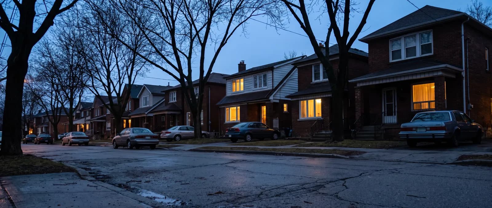 An ordinary residential street at dusk: modest houses along the pavement, a few windows lit warm from inside, bare trees and parked cars under a deep blue sky.