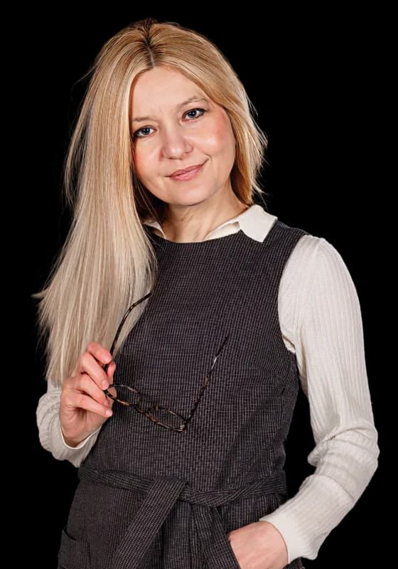 A teacher with long blonde hair in a dark pinafore, holding her glasses.