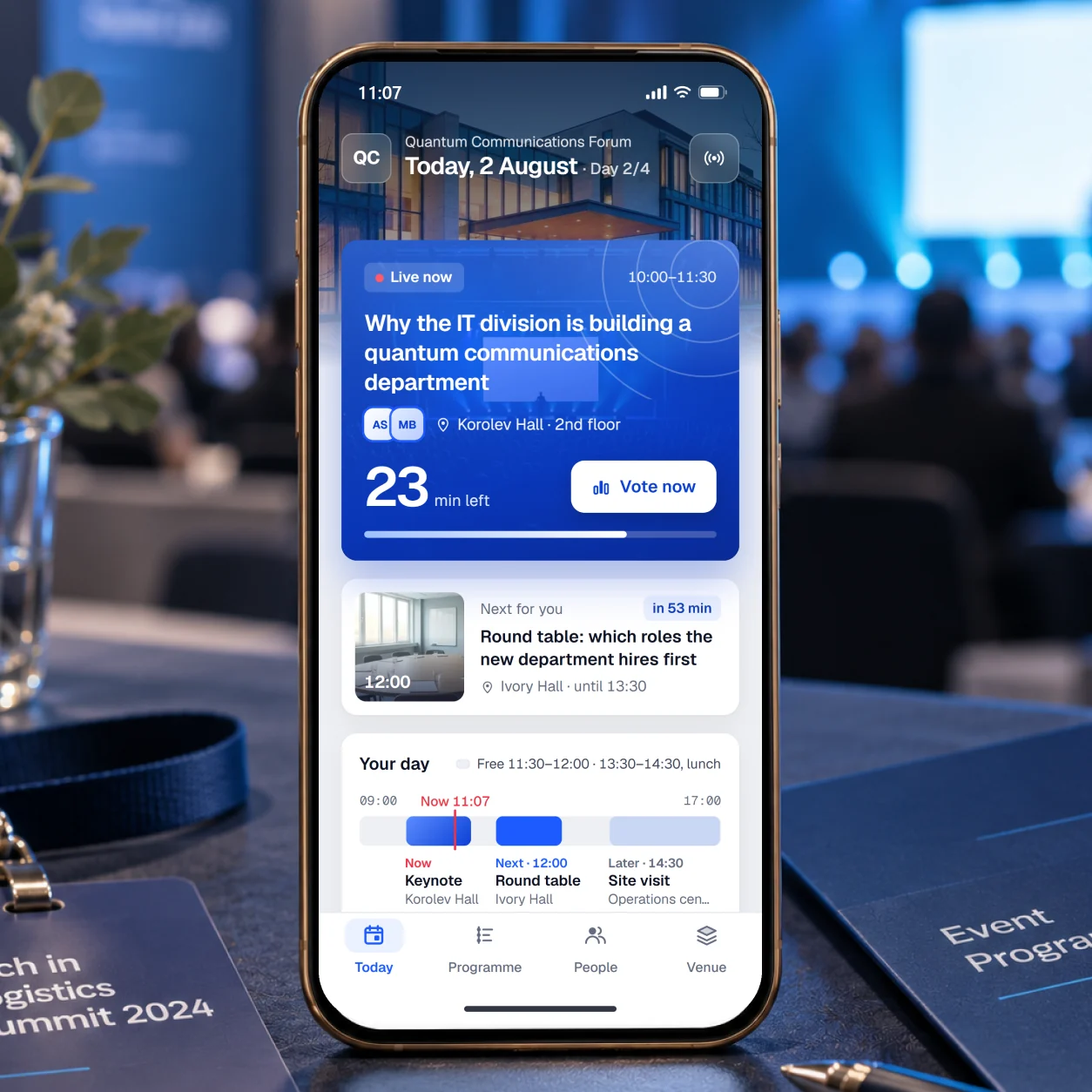 The events app on a phone at a conference: Today at the Quantum Communications Forum, the keynote live with 23 minutes left and a vote open, the round table next at 12:00, and the participant's day as a strip.