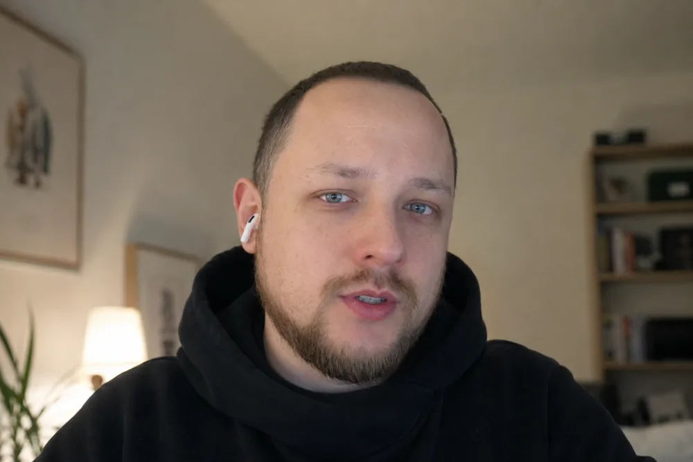 A man in a black hoodie with one earbud in, mid-sentence on a video call from a room with framed prints and a bookshelf.